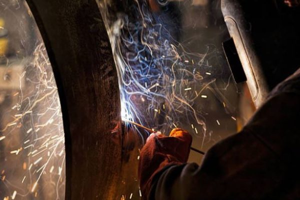 Welder in protective gear fusing metal, sparks flying around a curved metal surface at a workshop.